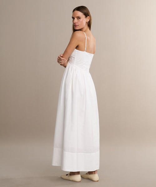 A woman in a white Peninsula Dress stands sideways, gazing over her shoulder. She pairs the dress with cream flats and is set against a plain, neutral background.