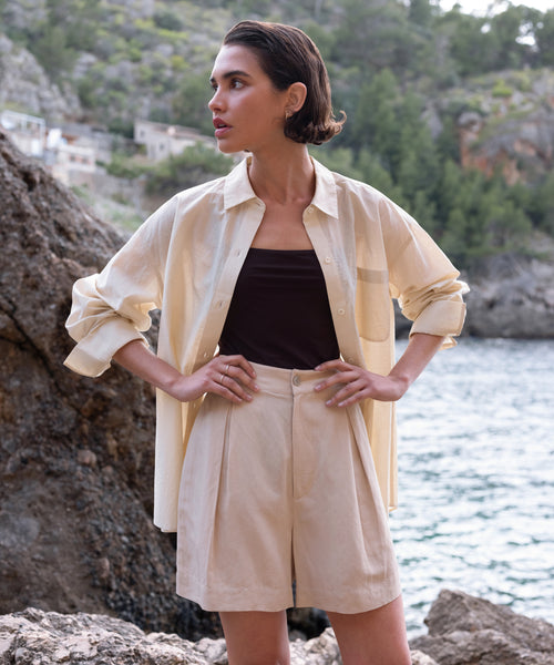 A woman stands outdoors by the water, wearing the Pierce Shirt in light beige over a dark top and matching shorts. The textured cotton Italian fabric shirt is paired with scenic rocky cliffs and trees in the background.