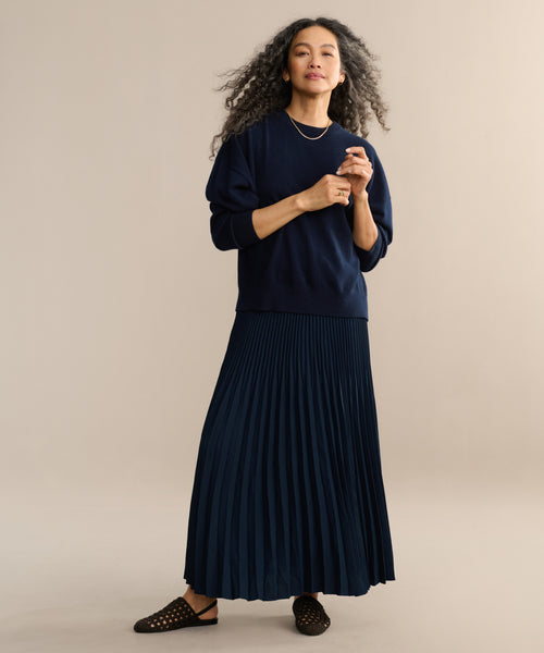 A woman with long curly hair wears the Pleated Skirt and a matching navy sweater, standing confidently against a beige background with gold jewelry and black slip-on shoes.