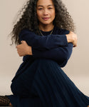 A woman with long, curly gray hair smiles softly while sitting on the floor in a Pleated Skirt and sweater, her arms wrapped around her knees against a plain background.