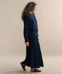 A smiling woman with curly hair models a pleated skirt and a navy sweater, walking barefoot on a beige background, facing left.