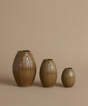 Three ribbed ceramic vases in graduated sizes with a glossy brown glaze, arranged against a solid beige background.