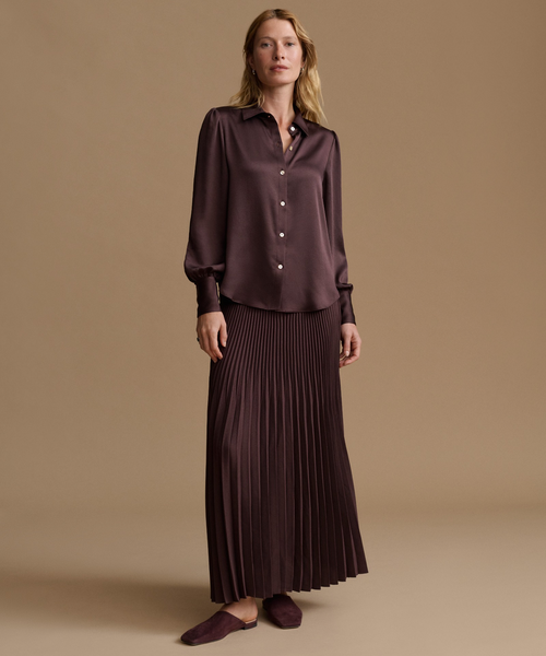 A woman stands in a studio wearing a dark plum satin button-front blouse with long sleeves and a matching pleated maxi skirt, paired with dark plum flats, against a warm beige background.