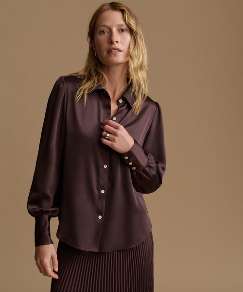 A woman modeling a dark plum satin button-up blouse with gold buttons, paired with a matching pleated skirt, against a solid tan studio background.