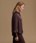 A woman in side profile wearing a dark plum satin blouse with balloon sleeves and a matching pleated skirt, set against a solid warm beige studio background.