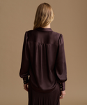 A woman viewed from behind wearing a dark plum satin blouse with long sleeves and gold-tone buttons on the cuffs, paired with a pleated skirt, against a solid tan studio background.