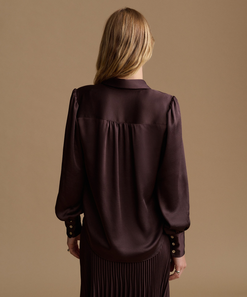 A woman viewed from behind wearing a dark plum satin blouse with long sleeves and gold-tone buttons on the cuffs, paired with a pleated skirt, against a solid tan studio background.