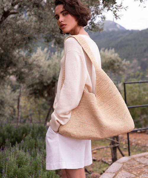 A person with short brown hair, in a cream sweater and white shorts, stands outdoors holding the Raffia Isla Bag. Greenery and mountains are visible in the background.