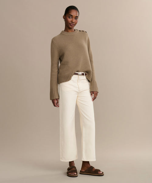 A woman poses against a beige background wearing the Ren jean in a wide-leg cream silhouette, paired with a tan sweater with button details, a brown belt, and brown sandals. Her pose is relaxed and her expression is neutral.