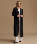 A woman stands against a warm beige studio background wearing a long black coat over a cream blouse and wide-leg cream trousers.