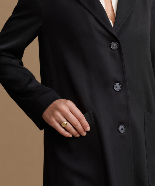 Close-up of a person wearing a black coat with dark buttons, a white shirt at the collar, and a gold ring on one finger, set against a solid tan background.
