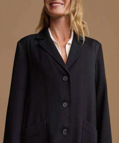 A close-up of a woman wearing a black blazer-style coat over a white collared shirt, against a warm beige studio background.