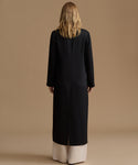 A woman viewed from behind wearing a long black coat over cream wide-leg pants, standing against a plain beige studio background.