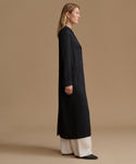 A woman stands in profile wearing a long black coat over cream wide-leg pants against a plain warm beige studio background.