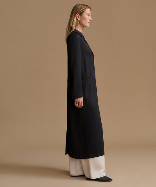 A woman stands in profile wearing a long black coat over cream wide-leg pants against a plain warm beige studio background.
