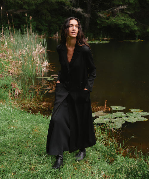 A woman stands outdoors by a lake wearing a long black coat over a dark outfit with black boots. Tall grasses, green grass, and lily pads are visible around the water, with trees in the background.