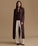 A woman stands against a plain beige studio background wearing a long dark plum coat over a matching dark plum top, paired with wide-leg cream pants and dark plum shoes.