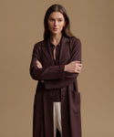 A woman modeling a long, dark plum-brown tailored coat over a matching top and white pants, standing against a solid warm beige studio background.