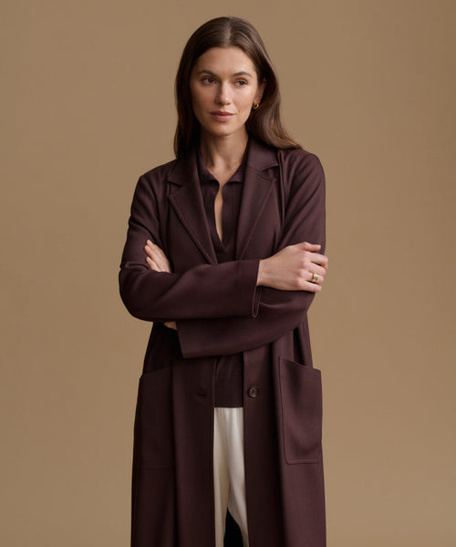 A woman modeling a long, dark plum-brown tailored coat over a matching top and white pants, standing against a solid warm beige studio background.
