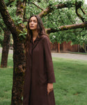 A woman wearing a long, dark brown collared coat stands outdoors in a grassy area next to a tree with mossy branches.