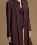A woman wearing a long, dark plum-brown coat over a matching top and light trousers, photographed against a plain warm beige studio background.