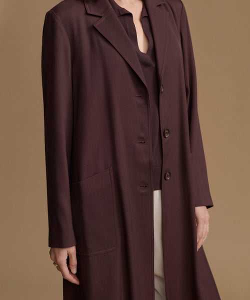 A woman wearing a long, dark plum-brown coat over a matching top and light trousers, photographed against a plain warm beige studio background.