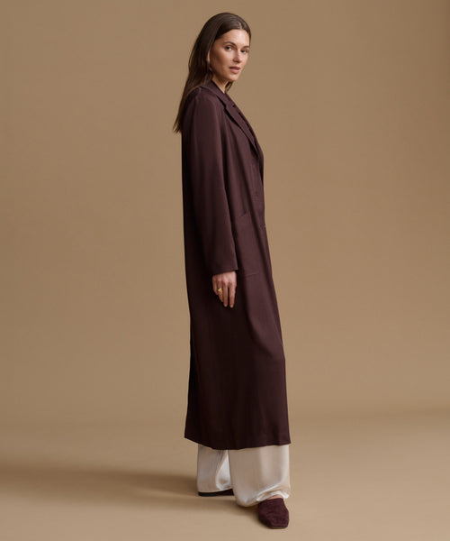 A woman stands in profile wearing a long, dark plum-brown coat over white wide-leg pants and matching dark slippers, photographed against a plain warm beige studio background.