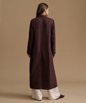 A woman viewed from behind wearing a long, dark plum-brown coat over cream wide-leg trousers, standing against a solid beige studio background.