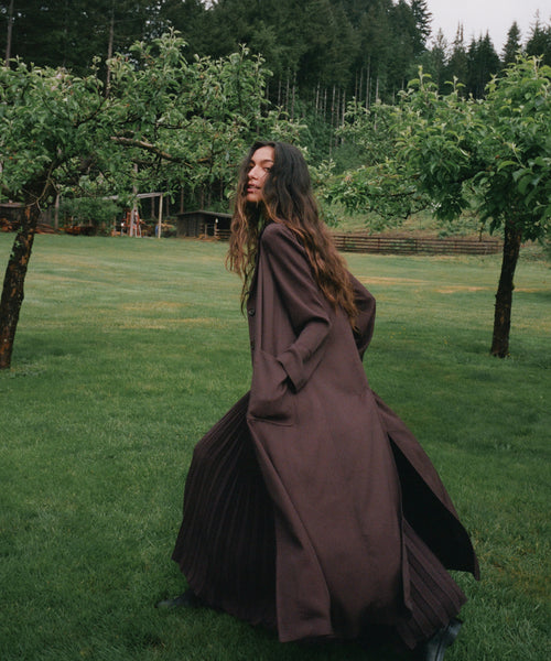 A woman wearing a long, dark brown flowing dress or robe stands on a grassy lawn with trees in the background.