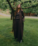A woman stands outdoors on a grassy lawn under a tree, wearing a dark brown long-sleeve collared coat over a matching dark brown top and pleated skirt, with black ankle boots.