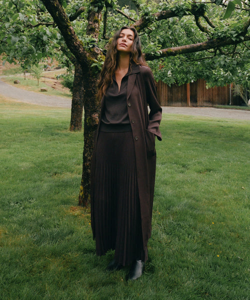 A woman stands outdoors on a grassy lawn under a tree, wearing a dark brown long-sleeve collared coat over a matching dark brown top and pleated skirt, with black ankle boots.