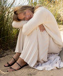 A blonde woman sits on sand, hugging her knees. She wears a cream sweater, long pleated skirt, and the Romy Sandal. Surrounded by tall grass, she smiles gently at the camera.
