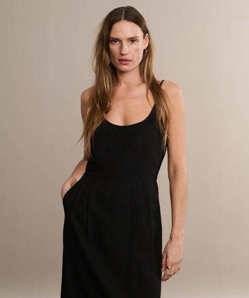 A woman with long brown hair wears the Rove Dress, standing against a plain beige background and looking at the camera with one hand in her pocket.