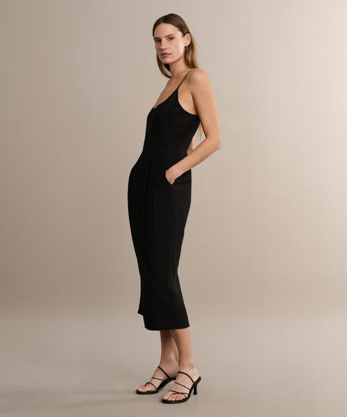 A woman poses in a neutral studio wearing the Rove Dress—a sleeveless black midi dress with thin straps—paired with black strappy heels. She faces the camera with her hands in her pockets.