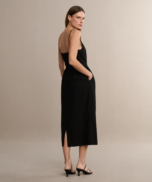 A woman stands on a light background wearing the sleeveless black Rove Dress, a midi dress with thin straps and side slit, styled with black heeled sandals. She poses with her hands in her pockets, glancing over her shoulder.