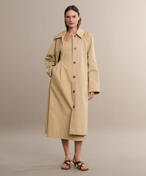 A woman stands against a plain background wearing the Rove Dress under a beige trench coat and brown sandals, with one hand in her pocket and a neutral expression.