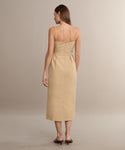 A woman with straight light brown hair faces away from the camera, wearing the Rove Dress—a khaki cotton spring dress with thin straps and a fitted waist—paired with brown heeled sandals against a neutral background.