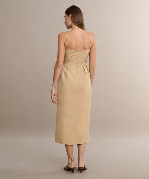 A woman with straight light brown hair faces away from the camera, wearing the Rove Dress—a khaki cotton spring dress with thin straps and a fitted waist—paired with brown heeled sandals against a neutral background.
