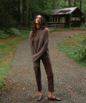 A woman stands on a gravel path in a wooded outdoor setting, wearing a ribbed brown mock-neck sweater and matching brown pants with brown loafers. A rustic cabin is visible in the background.