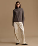 A woman stands against a warm beige studio background wearing a dark brown ribbed turtleneck sweater and cream wide-leg trousers.
