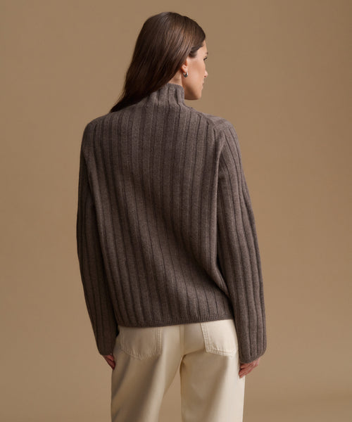 A woman shown from the back wearing a ribbed knit mock-neck sweater in a dark taupe-brown color, paired with cream-colored trousers, against a solid warm beige studio background.
