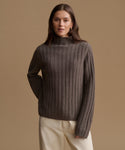 A woman wearing a ribbed knit mock neck sweater in a dark taupe-brown color, paired with cream trousers, set against a solid warm beige studio background.