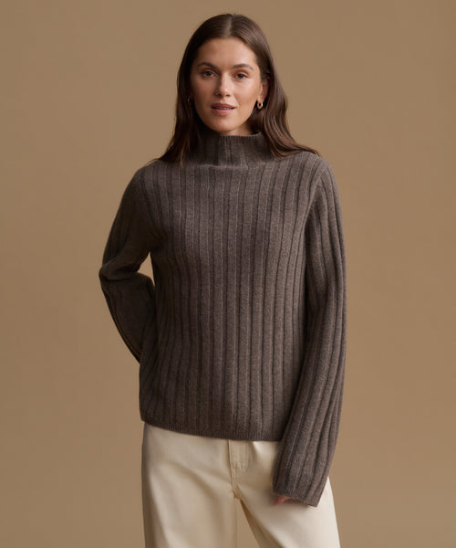 A woman wearing a ribbed knit mock neck sweater in a dark taupe-brown color, paired with cream trousers, set against a solid warm beige studio background.