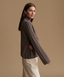A woman wearing a ribbed knit turtleneck sweater in taupe brown, paired with cream-colored trousers, posed against a solid warm beige studio background.