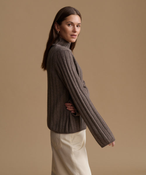 A woman wearing a ribbed knit turtleneck sweater in taupe brown, paired with cream-colored trousers, posed against a solid warm beige studio background.
