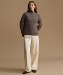 A woman stands against a warm beige studio background wearing a ribbed knit mock-neck sweater in dark taupe-brown, cream wide-leg trousers, and dark slip-on shoes.