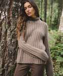 A woman wearing a ribbed knit mock neck sweater in taupe brown, leaning against a large tree trunk in a forest setting.