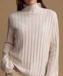A close-up of a person wearing a cream-colored ribbed knit mock neck sweater against a solid warm beige background.