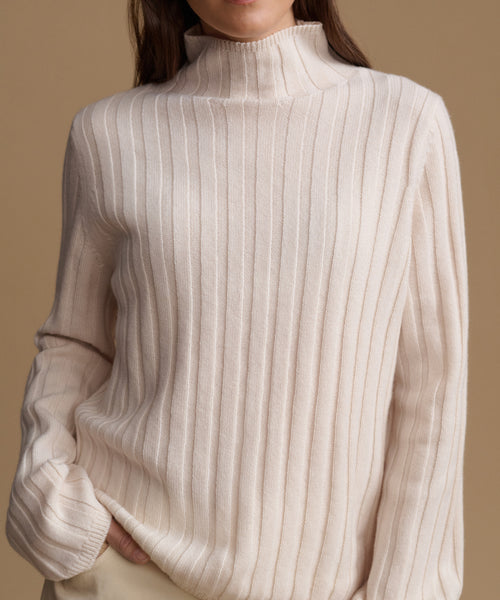 A close-up of a person wearing a cream-colored ribbed knit mock neck sweater against a solid warm beige background.