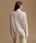 A woman shown from the back wearing a cream ribbed knit mock-neck sweater and matching cream trousers, set against a solid warm beige studio background.